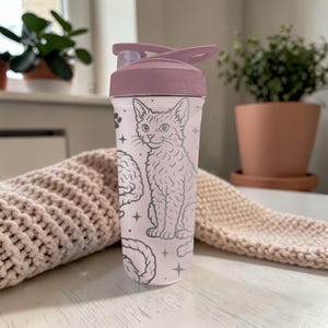 May include: A clear water bottle with a mauve lid and a cat illustration. The bottle features a design of a cat, brain, and stars in a light gray color. The bottle is sitting on a white surface.