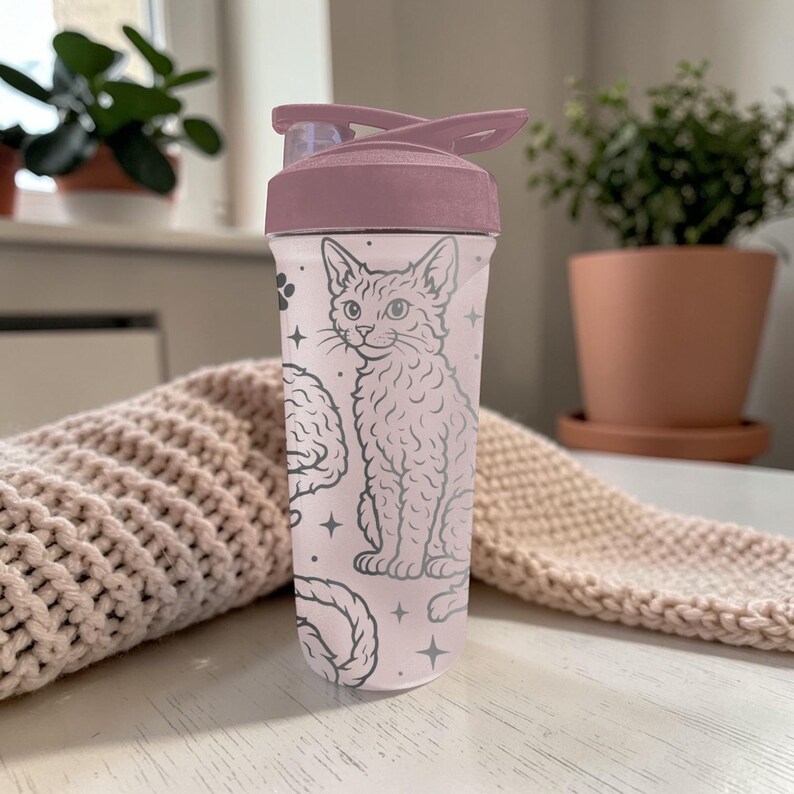 May include: A clear water bottle with a mauve lid and a cat illustration. The bottle features a design of a cat, brain, and stars in a light gray color. The bottle is sitting on a white surface.