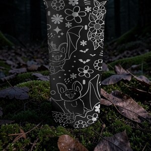 Pu&ograve; includere: Bicchiere nero con un motivo floreale e pipistrelli bianchi. Il design include vari fiori, stelle e illustrazioni di pipistrelli. Il bicchiere cilindrico &egrave; mostrato in posizione verticale su uno sfondo naturale.