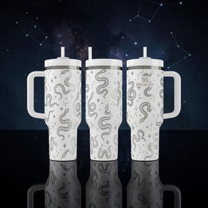 Könnte beinhalten: Ein weißer Stanley-Becher mit Henkel und Strohhalm, verziert mit einem sich wiederholenden schwarzen Linienmuster aus Schlangen und Sternbildern. Der Markenname Stanley ist in kleinen schwarzen Buchstaben oben auf dem Becher aufgedruckt.
