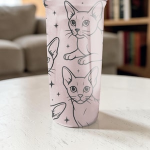 Puede incluir: Un vaso rosa con un patr&oacute;n repetido de caras de gatos negros y estrellas. Los gatos tienen orejas grandes y ojos almendrados. El vaso cil&iacute;ndrico es probablemente de pl&aacute;stico. Altura aproximada de 15 cm.