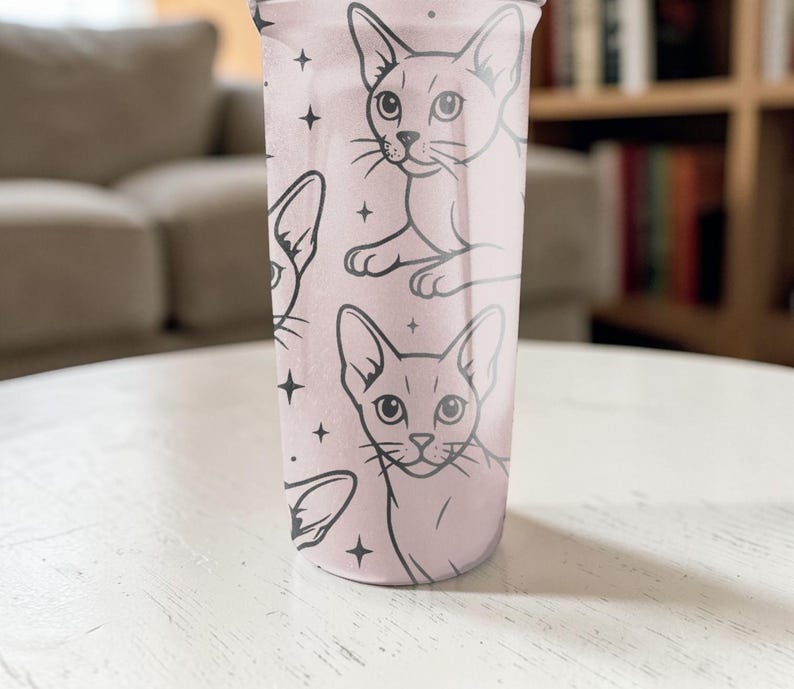 Puede incluir: Un vaso rosa con un patr&oacute;n repetido de caras de gatos negros y estrellas. Los gatos tienen orejas grandes y ojos almendrados. El vaso cil&iacute;ndrico es probablemente de pl&aacute;stico. Altura aproximada de 15 cm.