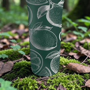 May include: A dark green tumbler with a white citrus fruit pattern. The design features detailed illustrations of whole and sliced citrus fruits, including oranges and lemons. The tumbler is cylindrical and stands upright against a natural, mossy background.