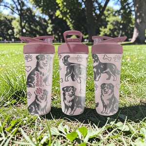 K&ouml;nnte beinhalten: Drei rosa Blender Bottles mit dunkelrosa Deckeln und Griffen, mit schwarz-wei&szlig;en Hundeillustrationen, Pfotenabdr&uuml;cken und Herzen. Auf den Flaschen steht "Blender Bottle". Die Flaschen stehen auf einer gr&uuml;nen, grasbewachsenen Oberfl&auml;che.