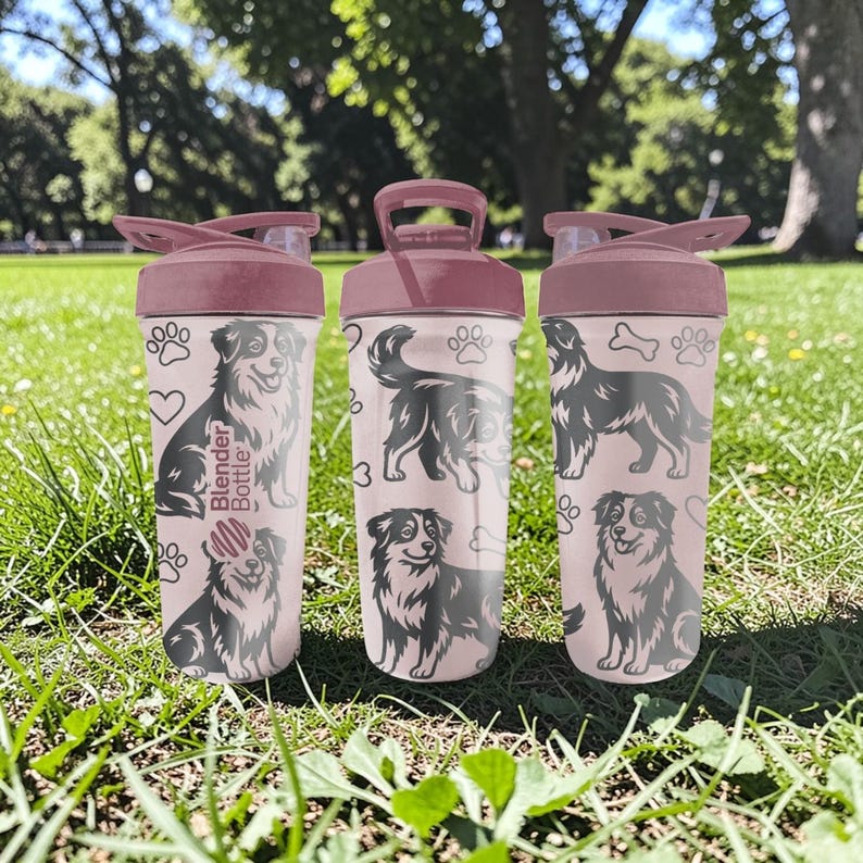 K&ouml;nnte beinhalten: Drei rosa Blender Bottles mit dunkelrosa Deckeln und Griffen, mit schwarz-wei&szlig;en Hundeillustrationen, Pfotenabdr&uuml;cken und Herzen. Auf den Flaschen steht "Blender Bottle". Die Flaschen stehen auf einer gr&uuml;nen, grasbewachsenen Oberfl&auml;che.