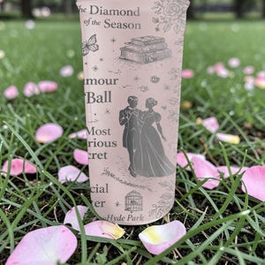 Pode incluir: Um copo alto e claro com texto e ilustra&ccedil;&otilde;es, incluindo um casal, livros e uma borboleta. O texto inclui "The Diamond of the Season", "amour Ball" e "Hyde Park". O copo &eacute; cercado por grama verde e p&eacute;talas de flores cor-de-rosa.