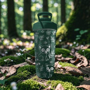 May include: A dark green water bottle with a handle and a screw-on lid. The bottle is decorated with white line art of lemurs, leaves, flowers, and stars. The background is a blurred forest scene with moss and leaves.