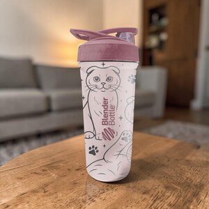 May include: A light pink Blender Bottle with a dark pink lid. The bottle features a cartoon cat illustration, paw prints, and the words "Blender Bottle". The bottle is sitting on a wooden table.