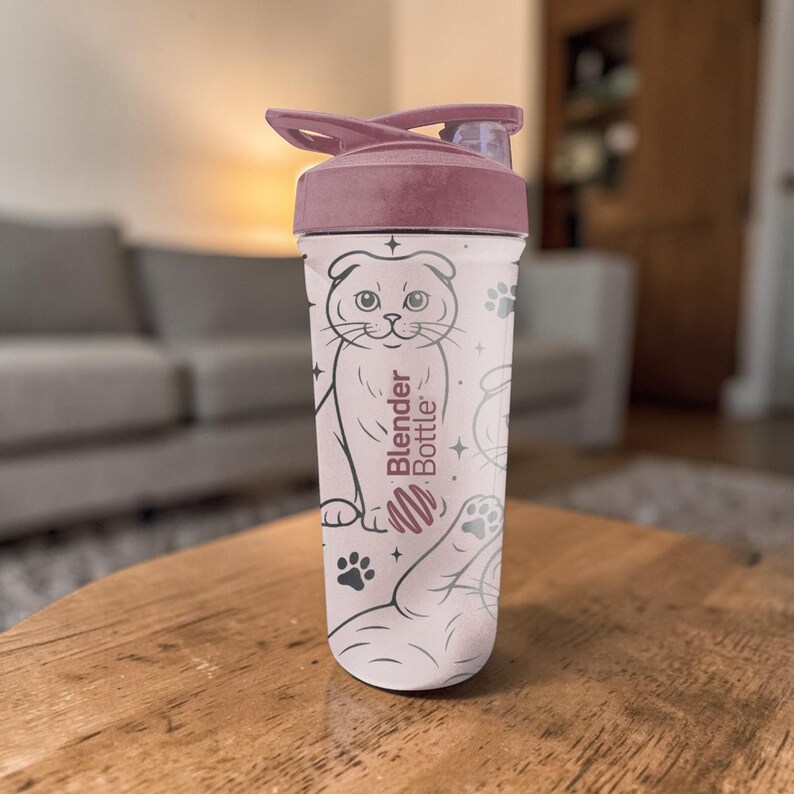 May include: A light pink Blender Bottle with a dark pink lid. The bottle features a cartoon cat illustration, paw prints, and the words "Blender Bottle". The bottle is sitting on a wooden table.