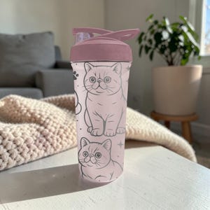 May include: A pink water bottle with a cat design. The bottle has a flip-top lid and a light pink body with a repeating pattern of cat illustrations and paw prints. The bottle is designed for hydration and is suitable for everyday use.
