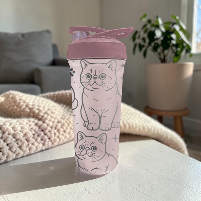 May include: A pink water bottle with a cat design. The bottle has a flip-top lid and a light pink body with a repeating pattern of cat illustrations and paw prints. The bottle is designed for hydration and is suitable for everyday use.