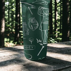 May include: Green tumbler with white line art of golf-related images, including a golfer, golf glove, golf ball, and tees. The word "DOGEY" is printed on the tumbler. The tumbler is on a wooden surface.
