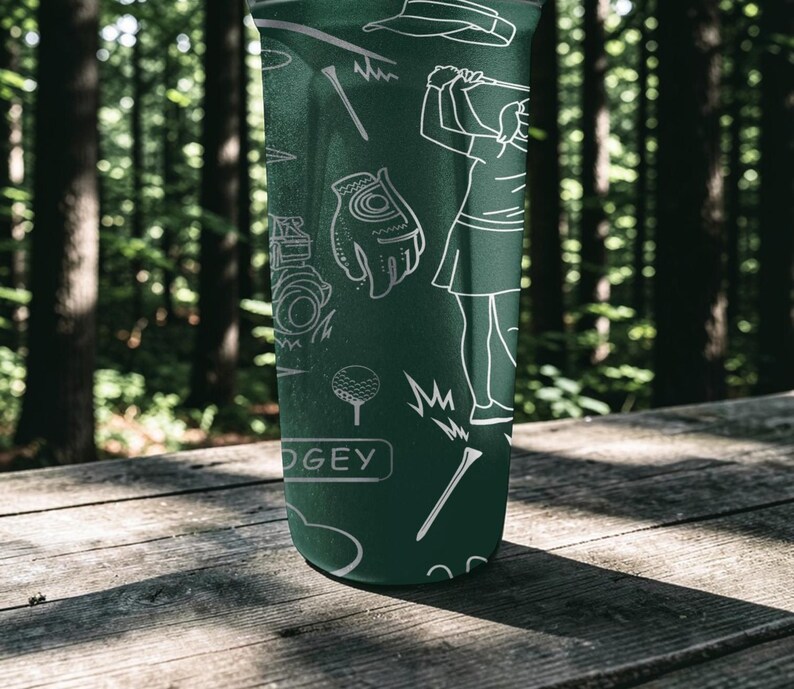May include: Green tumbler with white line art of golf-related images, including a golfer, golf glove, golf ball, and tees. The word "DOGEY" is printed on the tumbler. The tumbler is on a wooden surface.