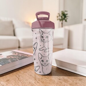 Puede incluir: Una botella de agua transparente con tapa y asa granate. La botella est&aacute; decorada con un patr&oacute;n repetido de gatos dibujados a l&iacute;nea y peque&ntilde;as estrellas. La botella es cil&iacute;ndrica y parece ser de pl&aacute;stico.