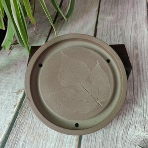 Op de afbeelding: Een ronde, lichtbruine keramische theebak met een bladontwerp. De bak heeft drie kleine gaatjes aan de onderkant.