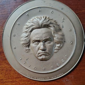May include: A bronze-coloured round medallion with a detailed relief portrait of Beethoven. The name "BEETHOVEN" is inscribed around the portrait, with the years "1770" and "1827" below. The text "MADE BY THE AUSTRIAN MINT, VIENNA, AUSTRIA" is at the bottom.