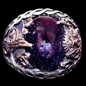 May include: A silver ring featuring a large, oval, deep blue stone with gold flecks. The ring has detailed silver and gold metalwork, with a braided border. The stone appears to be a type of aventurine.