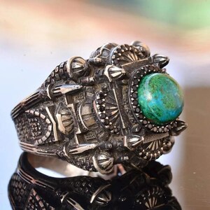 May include: A detailed silver ring with an intricate design. The ring features a large, round, green and blue stone at the center, surrounded by black stones. The band has a textured, industrial-inspired pattern.