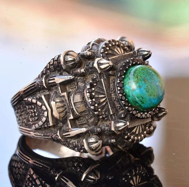 May include: A detailed silver ring with an intricate design. The ring features a large, round, green and blue stone at the center, surrounded by black stones. The band has a textured, industrial-inspired pattern.