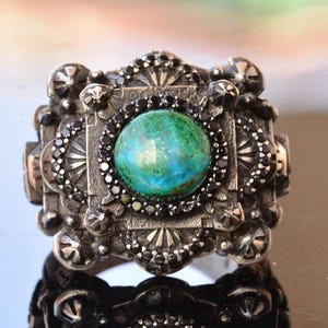 May include: A silver-toned ring with a large, round, turquoise-colored stone at its center. The ring is intricately designed with black accents and decorative patterns, showcasing a vintage or antique style.
