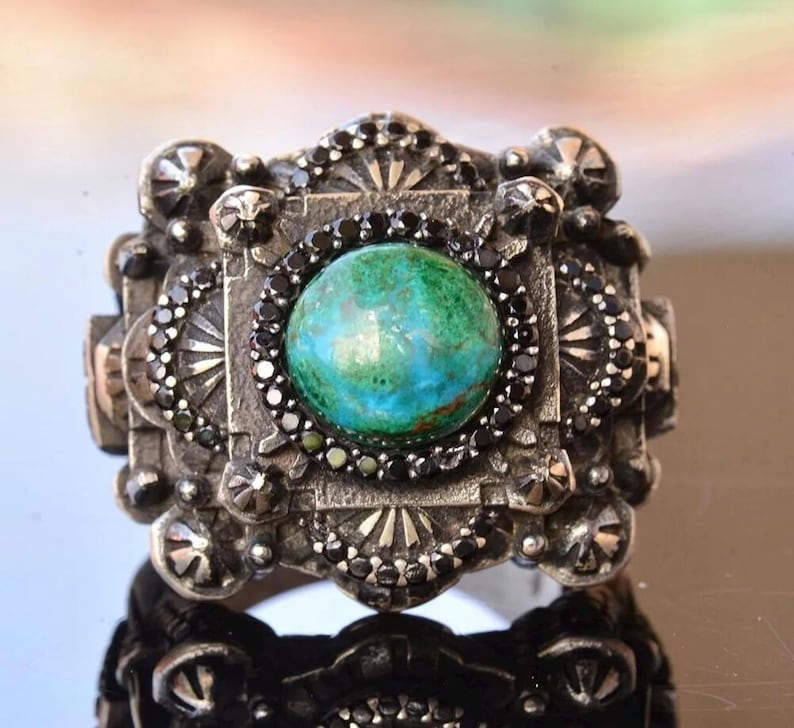 May include: A silver-toned ring with a large, round, turquoise-colored stone at its center. The ring is intricately designed with black accents and decorative patterns, showcasing a vintage or antique style.