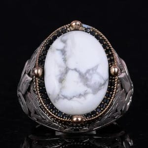 May include: A silver ring with an oval white stone, with grey veining. The stone is framed by a gold and black beaded border, and the ring band has a detailed leaf design. The ring is set against a dark background.