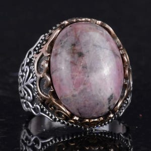May include: A silver ring featuring an oval, pink gemstone. The gemstone is set in a gold-coloured bezel. The ring has an ornate, filigree design on the band. The ring is displayed on a reflective black surface.