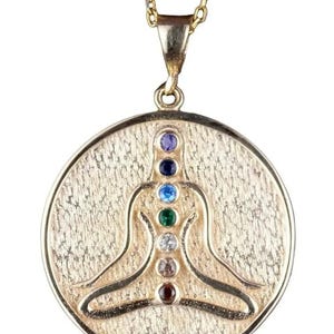 Könnte beinhalten: Goldfarbene Halskette mit Anhänger, die eine sitzende Figur mit sieben mehrfarbigen Edelsteinen darstellt, die Chakren repräsentieren. Der Anhänger hat eine strukturierte Oberfläche und hängt an einer zarten Goldkette. Ein spirituelles oder meditationsbezogenes Accessoire.