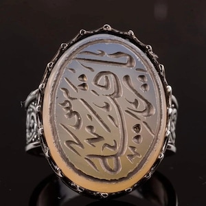 May include: A silver ring featuring an oval, pale stone with detailed calligraphy. The ring has an ornate band. The background is dark, highlighting the ring's design.