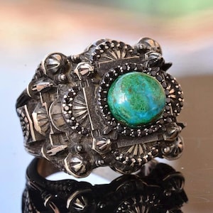 May include: A silver ring with an ornate design. The ring features a large, round, green and blue stone in the center, surrounded by black stones. The ring has a detailed, textured band with decorative elements.