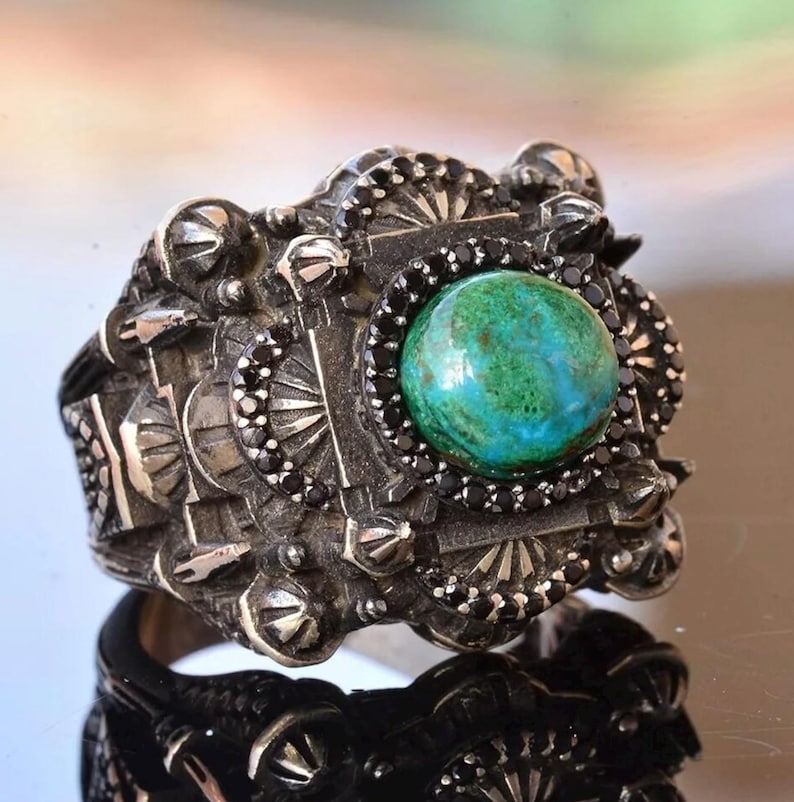 May include: A silver ring with an ornate design. The ring features a large, round, green and blue stone in the center, surrounded by black stones. The ring has a detailed, textured band with decorative elements.