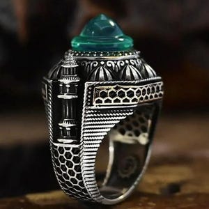 Puede incluir: Un anillo de plata detallado con una piedra preciosa de color turquesa. El anillo presenta intrincados diseños arquitectónicos, incluyendo una estructura similar a un minarete y un patrón de panal. La piedra preciosa está facetada.
