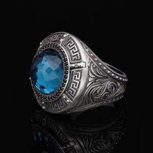 May include: A silver ring with an oval, faceted, light blue gemstone. The ring has a detailed, engraved design around the band and the gemstone setting, with a black border around the gemstone. The ring is displayed on a black surface.