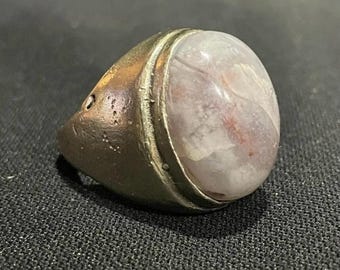 May include: A handcrafted ring featuring a large, oval, translucent stone with white and reddish tones. The ring has a tarnished bronze-coloured band, giving it a vintage look. The stone is set in a silver-coloured bezel.