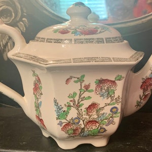 May include: A white porcelain teapot with a floral pattern in shades of pink, green, and blue. The teapot has a octagonal body and a lid with a knob on top.