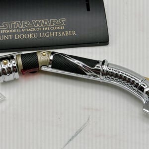 Puede incluir: Un sable de luz Count Dooku de Star Wars: Episodio II - El ataque de los clones. El sable de luz tiene un dise&ntilde;o plateado, negro y dorado con un mango curvo. Un soporte de exhibici&oacute;n negro con el t&iacute;tulo de la pel&iacute;cula est&aacute; detr&aacute;s del sable de luz.