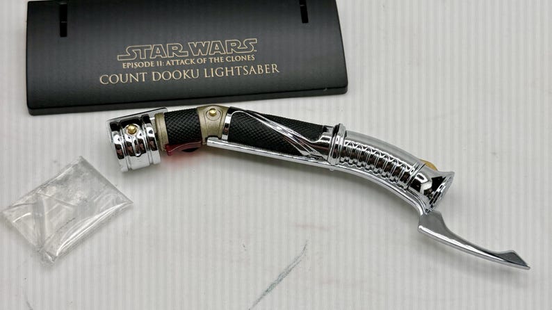 Puede incluir: Un sable de luz Count Dooku de Star Wars: Episodio II - El ataque de los clones. El sable de luz tiene un dise&ntilde;o plateado, negro y dorado con un mango curvo. Un soporte de exhibici&oacute;n negro con el t&iacute;tulo de la pel&iacute;cula est&aacute; detr&aacute;s del sable de luz.