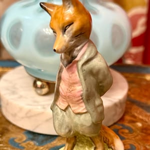 May include: A ceramic fox figurine standing on a small green base. The fox is anthropomorphic, wearing a light gray jacket, a pale pink vest, and matching light gray breeches. The fox has orange and white fur, pointed ears, and a long tail.