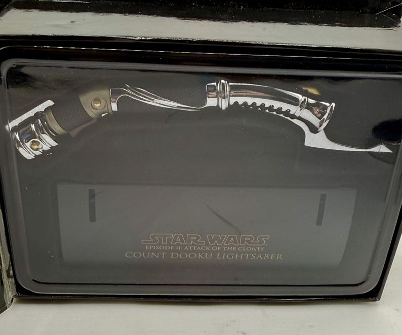 Puede incluir: Un sable de luz cromado y negro, presentado en una caja negra. El sable de luz tiene un mango curvo y detalles intrincados. El texto "STAR WARS EPISODE II: ATTACK OF THE CLONES COUNT DOOKU LIGHTSABER" es visible.