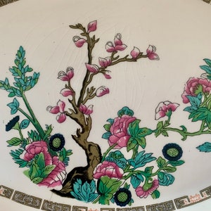 May include: A white ceramic plate with a floral design. The design features pink and green flowers, blue leaves, and a brown tree branch.