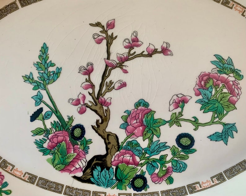 May include: A white ceramic plate with a floral design. The design features pink and green flowers, blue leaves, and a brown tree branch.