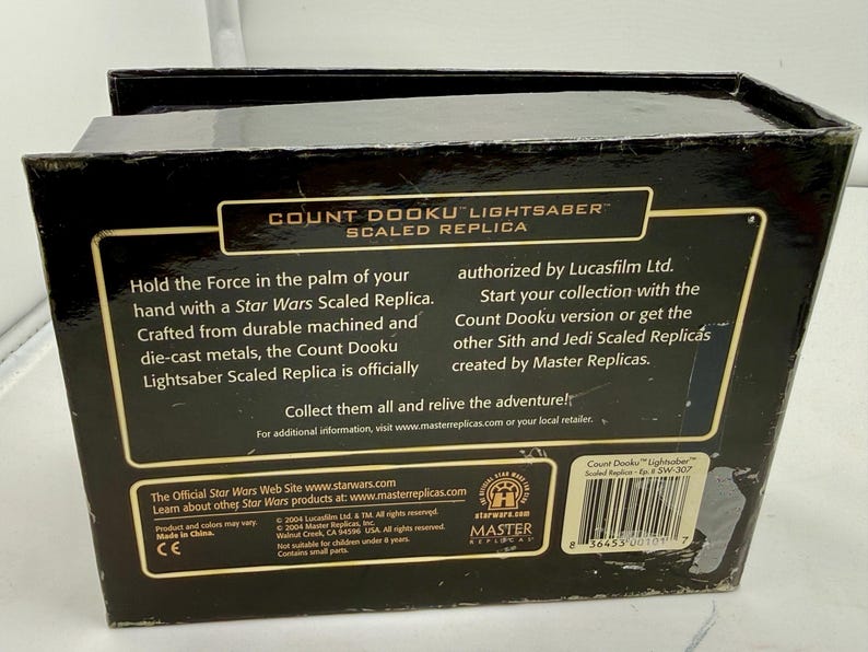Puede incluir: Una caja negra con el texto "COUNT DOOKU LIGHTSABER SCALED REPLICA" y la marca Star Wars. La caja describe la r&eacute;plica como fabricada con metales mecanizados y fundidos a presi&oacute;n duraderos. La caja tambi&eacute;n incluye un c&oacute;digo de barras y el texto "Made in China."