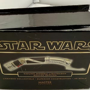 Puede incluir: Una caja negra con el logo de Star Wars en dorado. La caja presenta una imagen de una r&eacute;plica del sable de luz Count Dooku. El texto en la caja dice "Licensed Collectible - Superior Construction - .45 Scale".
