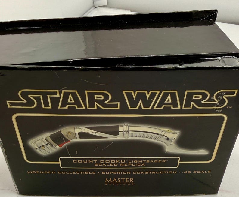 Puede incluir: Una caja negra con el logo de Star Wars en dorado. La caja presenta una imagen de una r&eacute;plica del sable de luz Count Dooku. El texto en la caja dice "Licensed Collectible - Superior Construction - .45 Scale".