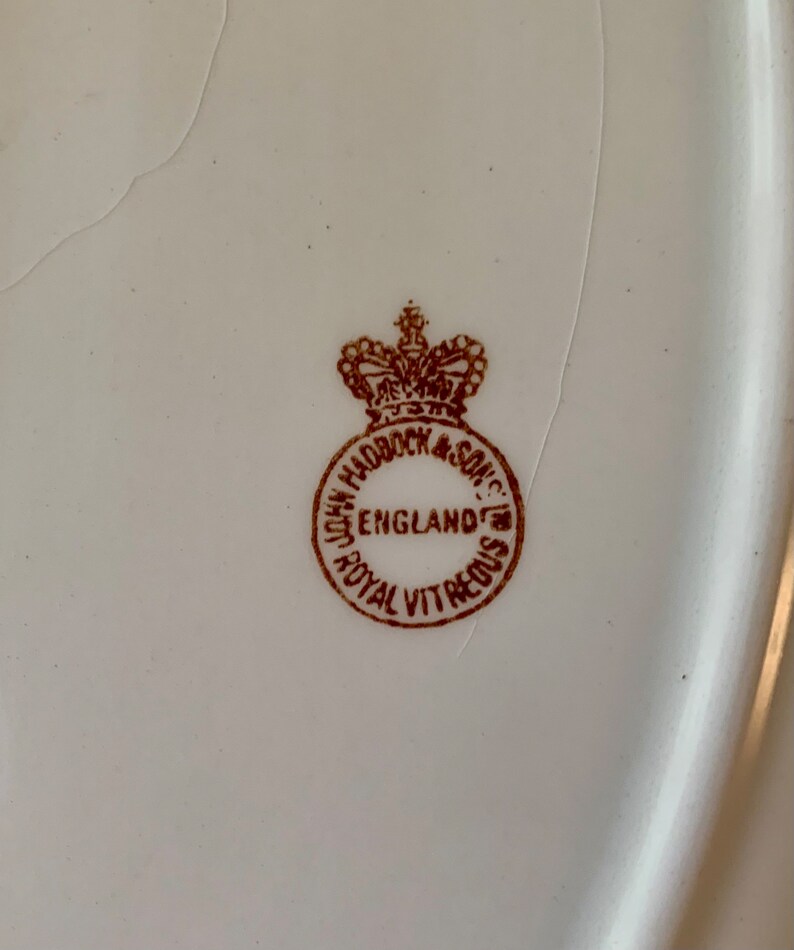 May include: Close-up of a brown stamp on a white ceramic plate. The stamp reads "John Haddock & Son, England, Royal Vitreous".