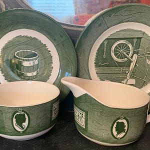 May include: A set of four green and white ceramic plates with a vintage design. The plates feature a green border with a white center. The center of each plate features a different design, including a bucket, a spinning wheel, and a floral pattern.