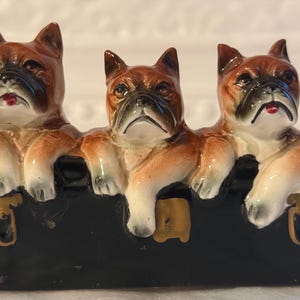 May include: A ceramic ornament depicting three Boxer dog heads emerging from a black trunk. The dogs have brown and white fur with black muzzles and a slightly grumpy expression. The trunk has gold detailing.