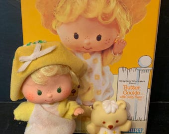 May include: A vintage Strawberry Shortcake doll set featuring a yellow-haired doll wearing a yellow and white dress and hat, a yellow teddy bear with a white bib, and a red comb. The box features the Strawberry Shortcake logo and the text "Welcome to the World of Strawberry Shortcake".