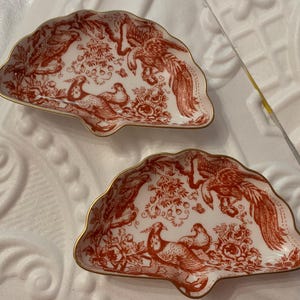 May include: Two fan-shaped decorative dishes. The dishes have a white background with a red bird and floral pattern. The edges are trimmed with gold. The dishes are likely made of ceramic or porcelain.
