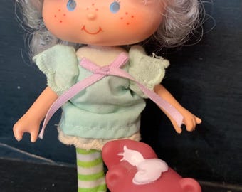May include: A vintage Strawberry Shortcake doll with white hair and a green dress. She is holding a pink and red plush bear with a white bib.